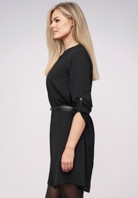 Blonde vrouw in een zwart lange mouwen jurk met opgerolde mouwen en een dunne zwarte riem, die naar links kijkt tegen een lichte achtergrond.