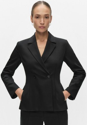 Femme avec les cheveux attachés portant un blazer noir à double boutonnage et un pantalon assorti, mains dans les poches, expression neutre, fond blanc.