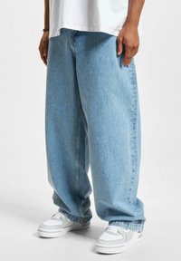 Lyseblå denim bukser med vide ben og avslappet passform, vist med hvite sneakers som har grå og lysegrønne detaljer.