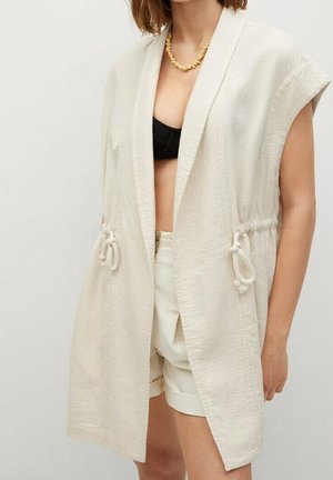 Ærmeløs beige tekstureret vest med snøre i taljen båret over sort top og beige højttaljede shorts på en person.