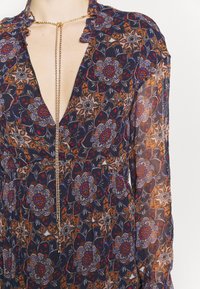 Robe fleurie bleu marine avec un décolleté en V profond, un col à volants, des manches longues en voile transparent et un détail de chaîne métallique. Comprend un tissu à motifs complexes.