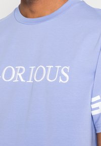 Camiseta de algodón de color azul claro con la palabra "GLORIOUS" en bordado blanco, con acentos de rayas blancas en las mangas.