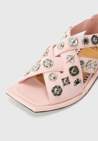 Pink sandal made of soft material with cross straps, adorned with various silver decorative studs and a square toe design.
