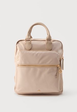 Sac à dos beige, de forme carrée, avec poignées supérieures, poche avant zippée et détails zippés dorés sur fond blanc.