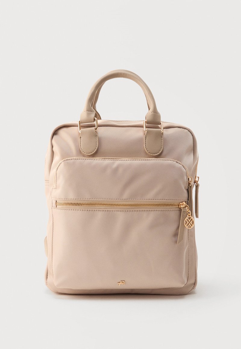 Sac à dos beige, de forme carrée, avec poignées supérieures, poche avant zippée et détails zippés dorés sur fond blanc.