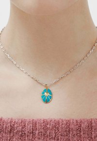 Collar de cadena de plata con un colgante ovalado de turquesa, detalle de corazón dorado y patrón geométrico. Diseñado para un ajuste ceñido alrededor del cuello.