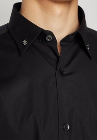 Nahaufnahme einer Person, die ein schwarzes Hemd mit Knöpfen trägt, an dessen Kragen Metallstifte mit der Aufschrift "HU" und einem kleinen Design befestigt sind.