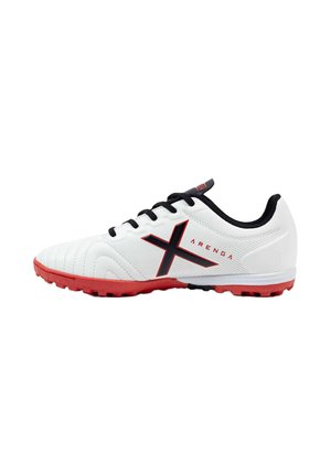 Zapatilla deportiva blanca con cordones negros, logo "X" en negro y rojo, suela roja y forro interior negro, diseñada para superficies de césped artificial o turf.