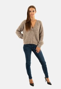 Cardigan in maglia beige con scollatura a V, texture a costine e chiusura con bottoni, abbinato a jeans scuri e tacchi alti neri. Look semplice e casual.