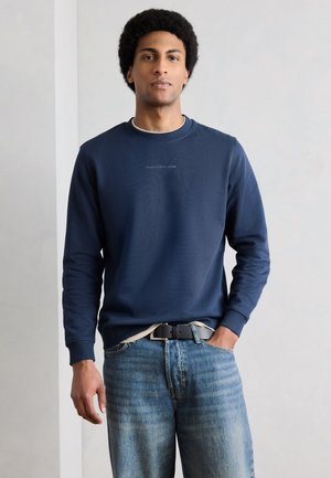 Jeune homme avec une coiffure afro portant un sweat-shirt bleu marine et un jean bleu, debout avec une main dans la poche devant un fond uni.
