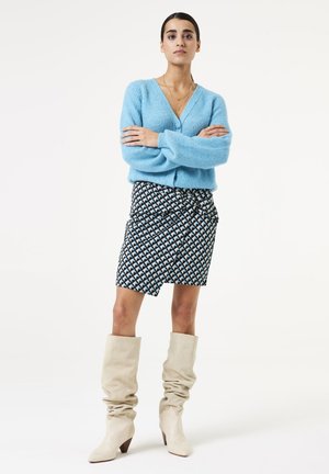 Cardigan en tricot côtelé bleu clair, jupe noire et blanche à motif avec un lien, bottes crèmes lâches jusqu'au genou, à bout pointu et avec un petit talon.