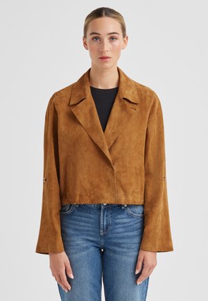 Frau mit zurückgebundenem Haar, die eine braune, kurz geschnittene Wildlederjacke über einem schwarzen Oberteil und blauen Jeans trägt, und vor einem schlichten grauen Hintergrund steht.