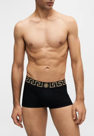 Buste masculin portant un boxer noir avec un motif géométrique doré et le logo Méduse sur la ceinture, debout sur un fond uni.