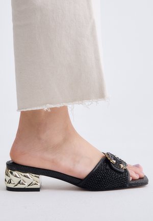 Pie con mule tejido negro con hebilla decorativa dorada y tacón bloque geométrico dorado, llevando pantalones beige claro con dobladillo deshilachado.