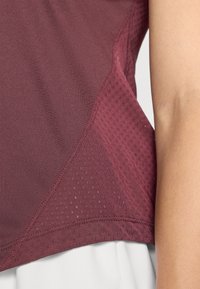 Bordeauxrotes Sportoberteil mit einem glatten Stoff, der Netzpaneele und abgestochene Kanten aufweist und Atmungsaktivität sowie Komfort betont.