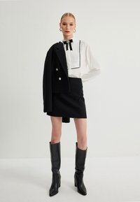 Conjunto de falda negro con una falda texturizada, blusa blanca con ribetes negros y un acento de lazo, combinada con botas negras hasta la rodilla.