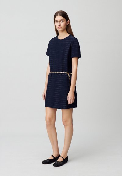 Vestido de manga corta azul marino con rayas horizontales, que cuenta con un cinturón de cadena dorada y dos bolsillos laterales; combinado con zapatos negros de tela trenzada sin cordones.