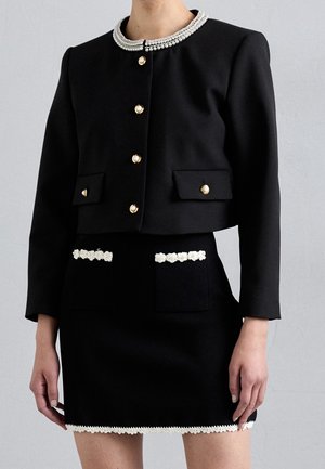 Veste courte noire avec boutons en perle et col, associée à une jupe noire assortie avec bordure florale blanche sur les poches et l'ourlet.