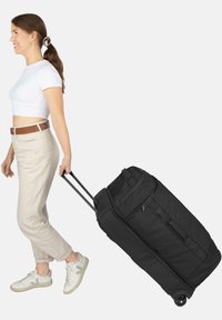 Schwarzer Rollkoffer mit zwei ausziehbaren Griffen, aus Stoffmaterial, großen Reißverschlussfächern und robusten Rädern, der von einer Person gezogen wird.