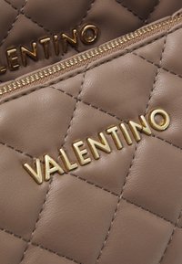 Borsetta in pelle trapuntata color taupe con dettagli in oro e logo "VALENTINO" ben visibile sulla superficie. Particolare della chiusura con cerniera visibile.