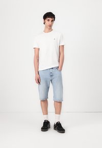 T-shirt blanche à manches courtes avec un petit logo, shorts en denim bleu clair, et baskets noires avec des accents blancs. Fond uni.