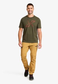 Olivgrön t-shirt med en röd grafisk logotyp, kombinerad med beige cargobyxor med dragkedjefickor och en avsmalnande passform. Svarta sportskor.