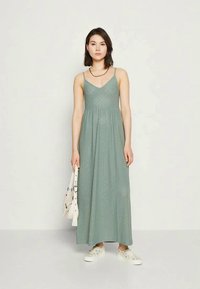 Maxi obleka v umirjeni zeleni barvi z tankimi naramnicami, teksturirano blago z geometrijskimi vzorci, v kombinaciji z belimi supergami in torbico.