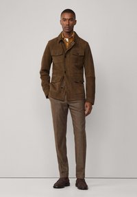 Braune Wildlederjacke mit zwei Brusttaschen, Knopfverschluß und Kragen; kombiniert mit braunen strukturierten Hosen und dunklen Schuhen.