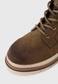 Brązowe zamszowe buty z teksturowaną cholewką, wzmocnionym czubkiem i beżową gumową podeszwą. Posiadają metalowe oczka i brązowe sznurowadła z wzorowanym splotem.