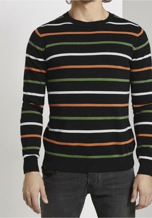 Mann trägt schwarzen Strickpullover mit horizontalen weißen, grünen und orangefarbenen Streifen, kombiniert mit dunkelgrauer Hose, steht vor neutralem Hintergrund.