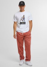 Bílý tričko s grafickým designem a textem, doplněné růžovými sportovními kalhotami s bílými bočními pruhy. Černá čepice dotváří celý outfit.