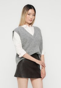 Młoda kobieta z długimi włosami w kolorze ombre, nosząca białą koszulę z podwiniętymi rękawami, szary sweter bez rękawów z dzianiny oraz czarną skórzaną mini spódniczkę.