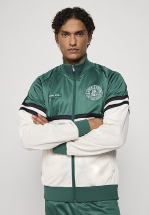Homme aux cheveux bouclés portant une veste de sport verte et blanche à zip, bras croisés, regardant droit devant sur un fond uni.