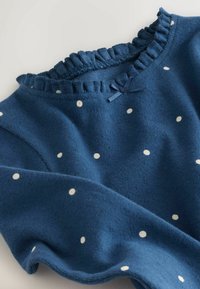 Blauwe trui met witte stippen, geribbelde halslijn, ruches aan de rand en een kleine blauwe strik als accent. Zachte textuur en een ontspannen pasvorm.