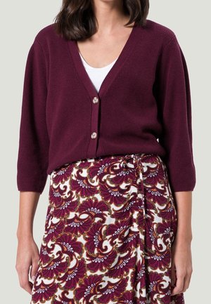 Frau trägt eine burgunderfarbene, V-förmige Strickjacke mit Knöpfen über einem weißen Oberteil und einem Rock mit einem Blumenmuster in Burgunderrot, Weiß und Gold.