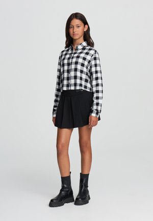 Camicia a quadri bianca e nera con maniche lunghe, abbinata a una gonna nera plissettata e stivaletti neri alla caviglia. Tessuti morbidi, design casual.