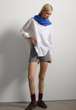 Weiße übergroße Bluse, graue gerippte Shorts, blauer gestrickter Schal, burgunderfarbene Socken und braune Schuhe. Glatter Stoff und sichtbare mehrschichtige Texturen.