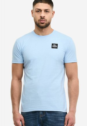 Moški, ki nosi svetlo modro kratko majico s črnim pravokotnim logotipom "Lonsdale London" na levi strani prsi.