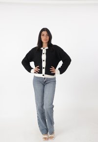Cardigan negro de punto con un collar blanco, que presenta grandes botones negros en la parte delantera, combinado con jeans azul claro. Textura suave.