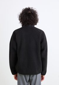 Sort fleece-pullover med en struktureret overflade, raglanærmer og høj krave, båret over grå bukser. Minimalistisk design uden synlige logoer.