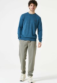 Man met krullend haar, gekleed in een blauwe trui, grijze broek met trekkoord en witte sneakers, staand voor een eenvoudige witte achtergrond.