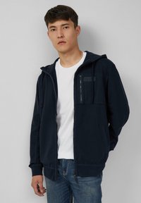 Marineblauwe zip-up hoodie met een voorzak en een borstrits, voorzien van een trekkoordcapuchon en geribbelde mouwen. Gedeeltelijk over een wit T-shirt gedragen.