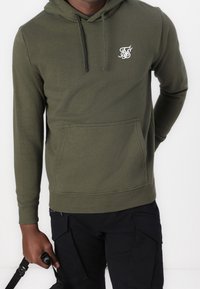 Sudadera de color verde oliva hecha de algodón, con un bolsillo tipo canguro en la parte frontal y capucha con cordón. Presenta un pequeño logo blanco en el pecho.