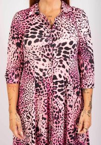 Rosa leopardmönstrad skjortklänning med trekvartsärmar, som har en slät textur och en utsvängd design. Accessoarer och tatueringar synliga.