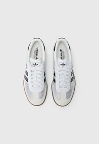 Scarpe sportive bianche con strisce nere, materiali in mesh e pelle, punta rotonda, suola in gomma e design testurizzato. Logo del marchio sulla linguetta.