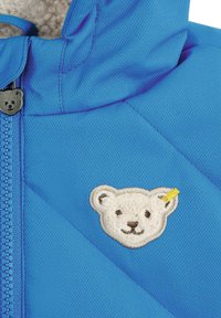 Blaue isolierte Jacke aus strukturiertem Stoff mit diagonalen Nähten und einem flauschigen Bärenpatch mit gelbem Akzent. Mit Reißverschluss.