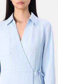 Abito avvolgente blu chiaro realizzato in tessuto testurizzato, caratterizzato da maniche lunghe, colletto e dettaglio di legatura laterale. Include una tasca sul petto.
