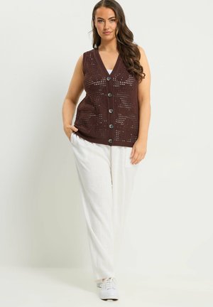 Vrouw draagt een donkerbruist knopjesgebreid mouwloos vest over een witte top, witte broek en witte sneakers, staand met één hand in haar zak.