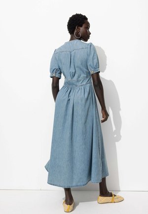 Robe maxi en denim avec des manches bouffantes, taille cintrée et jupe froncée. Couleur bleu clair, accents de boutons et tissu texturé. Chaussures plates jaunes.