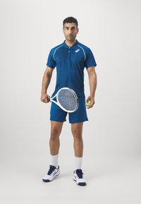 Sportlane, kes kannab tumesinist polo särki laimikroheliste aktsentidega, sobivaid lühikesi pükse, hoiab tennisereketit ja palli ning kannab valgeid sokke ja jalanõusid.